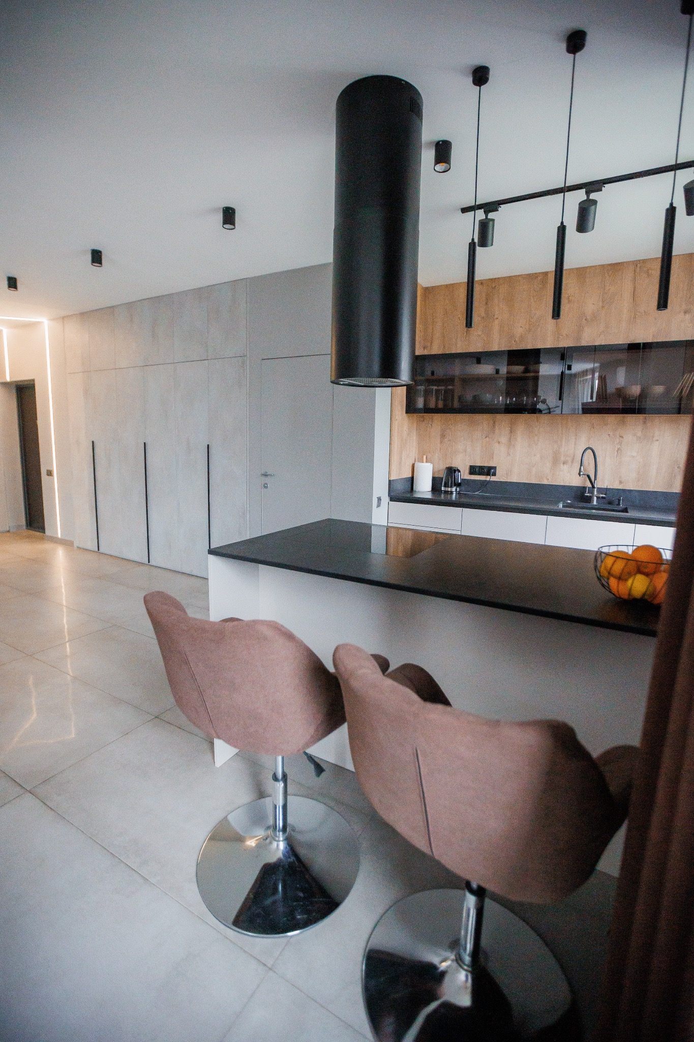 Nowoczesna kuchnia z wyspą i hokerami, czarny okrągły okap nad wyspą, oświetlenie szynowe i szafki z drewnianymi frontami w tle.