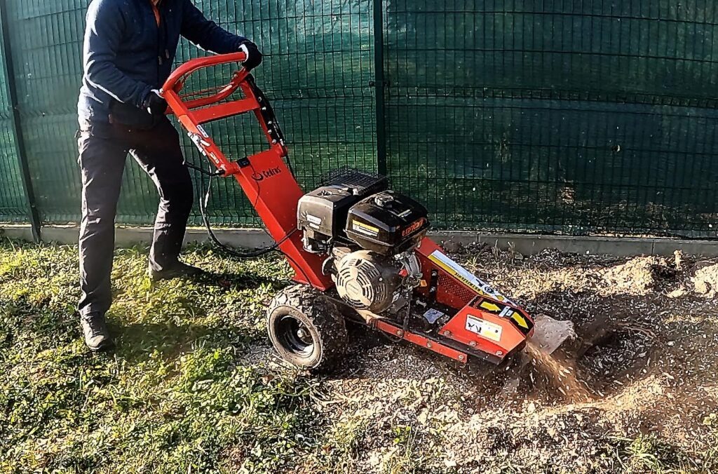 Operator frezuje pień drzewa pomarańczową frezarką spalinową na trawniku, z zielonym ogrodzeniem w tle. Wokół frezarki widać odłamki drewna i ziemi.