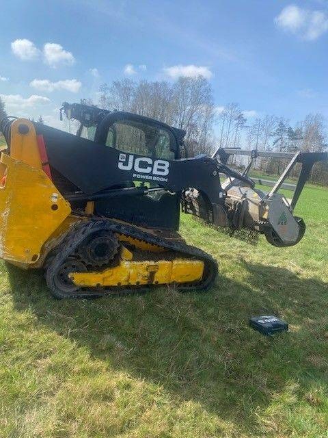 Żółto-czarna frezarka do pni JCB na gąsienicach, stojąca na trawie. Frezarka z zamontowaną głowicą frezującą. W tle drzewa i niebo z chmurami.