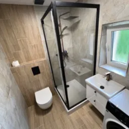 Wyremontowana łazienka z kabiną prysznicową typu walk-in, umywalką z szafką i toaletą podwieszaną. Płytki imitujące drewno na podłodze i ścianach, okno z widokiem na zieleń.