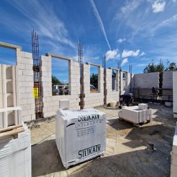 Mariusz Hałas - Wznoszenie ścian budynku z bloczków silikatowych, widoczne zbrojenia w otworach okiennych, palety z zapakowanymi bloczkami na placu budowy, pracownik w tle.