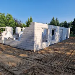 Wznoszenie ścian parteru domu z białych bloczków, widoczne ślady opon na piaszczystym gruncie, w tle zieleń drzew i błękitne niebo.