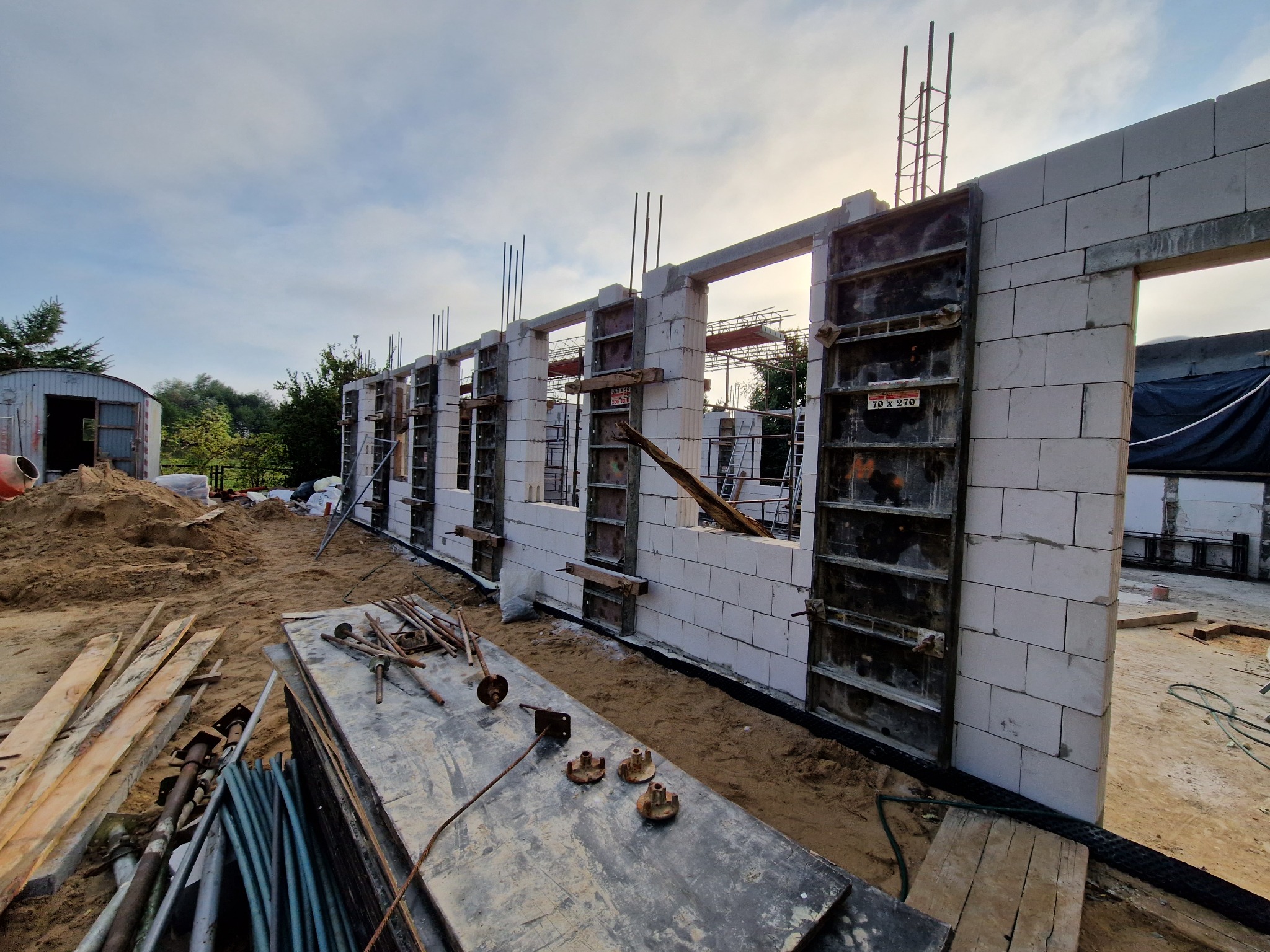 Budowa domu w trakcie: wznoszone ściany fundamentowe z bloczków betonowych z szalunkami, widoczne zbrojenia pionowe, piasek i narzędzia budowlane na pierwszym planie, pochmurne niebo w tle.