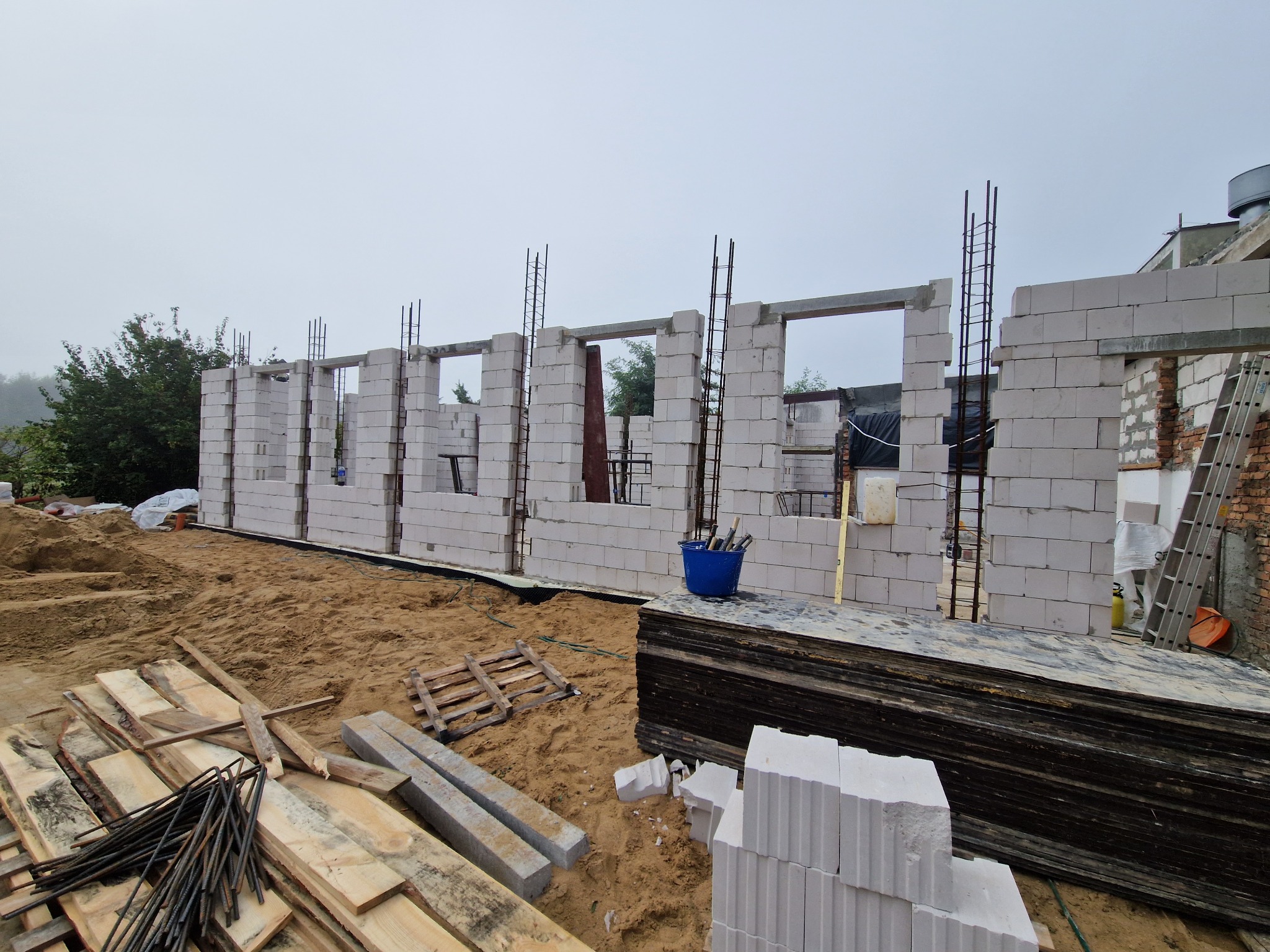 Wznoszenie ścian parteru budynku z betonu komórkowego Ytong, zbrojone pionowymi prętami stalowymi, widoczne deski szalunkowe i narzędzia na placu budowy.