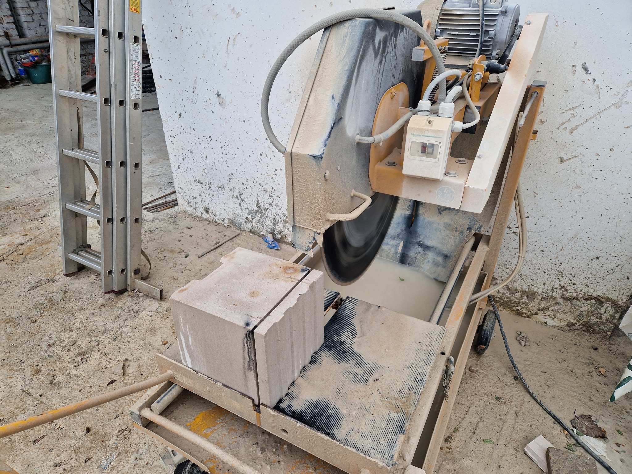 Piła do cięcia bloczków fundamentowych w trakcie pracy, z widocznym mokrym cięciem i aluminiową drabiną w tle.