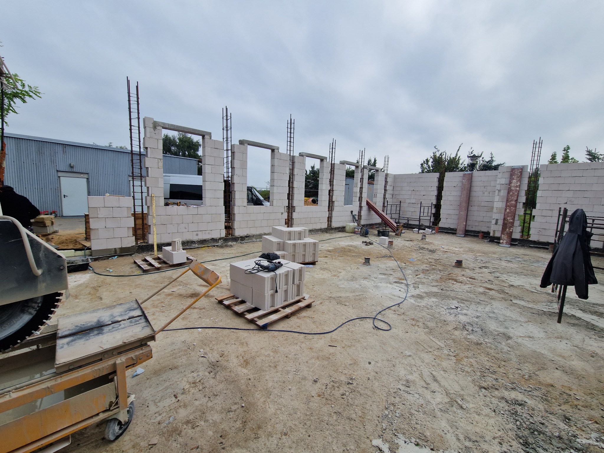 Wznoszenie ścian fundamentowych z betonu komórkowego Ytong, widoczne zbrojenie pionowe, paleta z bloczkami i przecinarka do betonu na placu budowy.