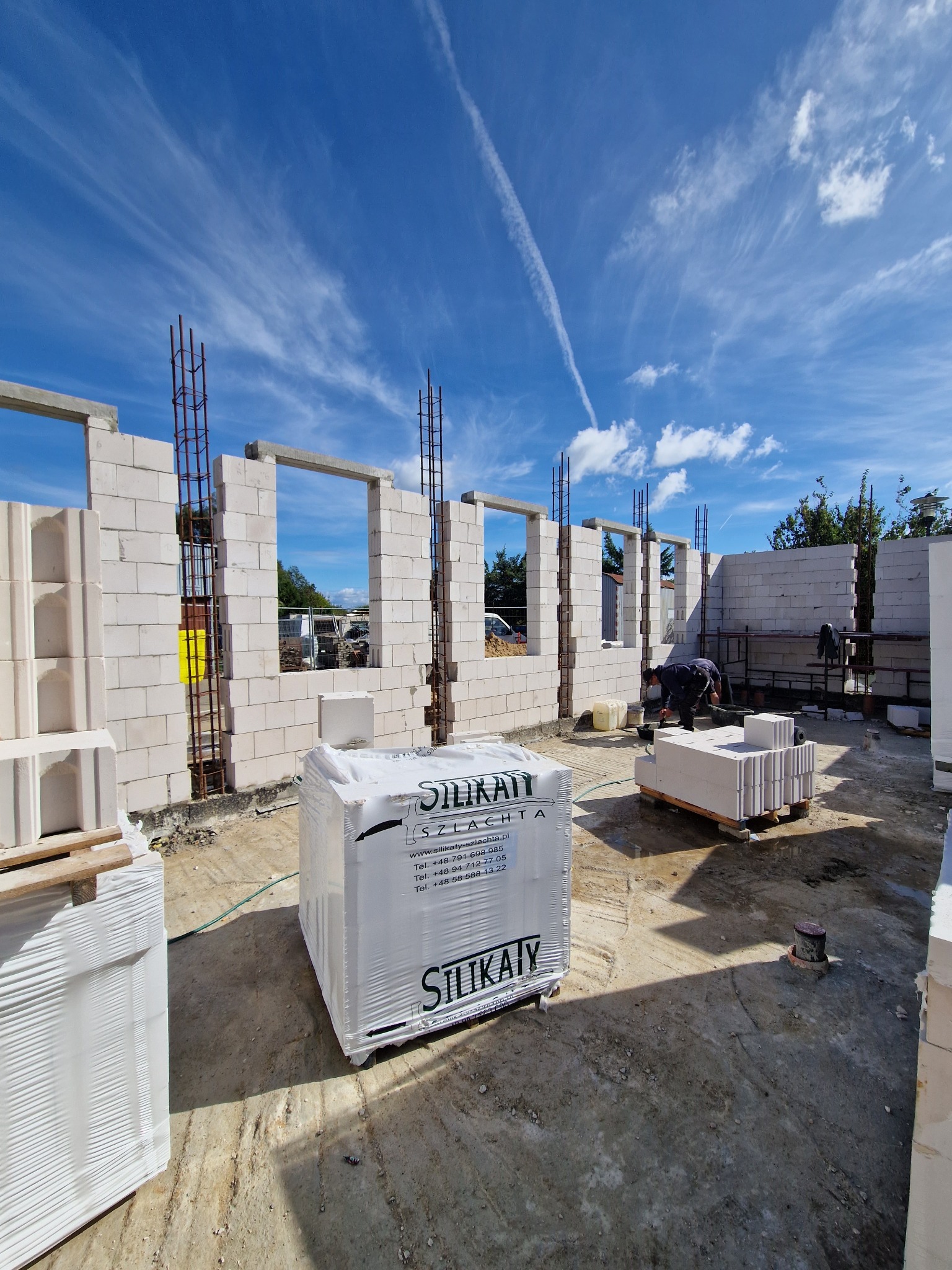 Wznoszenie ścian budynku z bloczków silikatowych, widoczne zbrojenia w otworach okiennych, palety z zapakowanymi bloczkami na placu budowy, pracownik w tle.