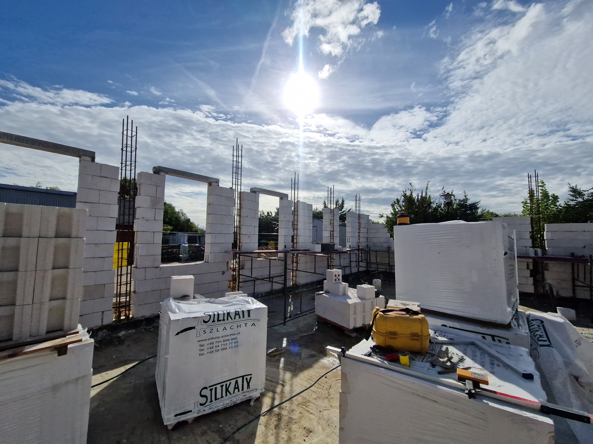 Wznoszenie ścian budynku z białych bloczków silikatowych, widoczne zbrojenia i palety z materiałem budowlanym, jasne słońce i błękitne niebo z chmurami w tle.