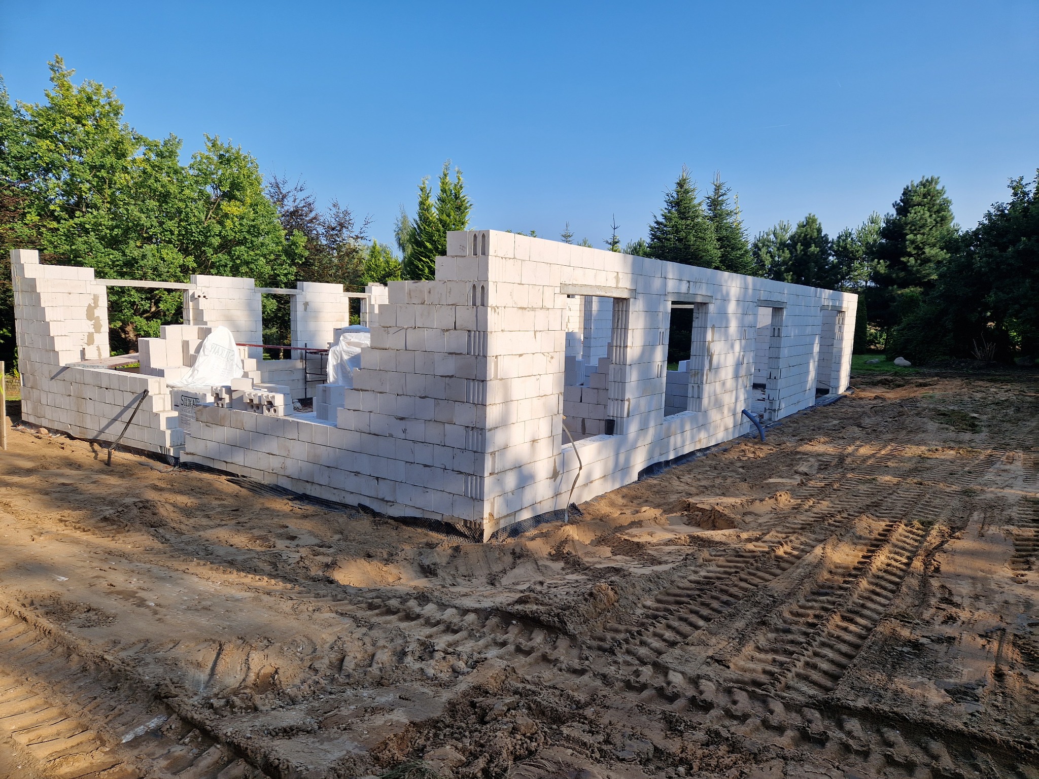 Wznoszenie ścian parteru domu z białych bloczków, widoczne ślady opon na piaszczystym gruncie, w tle zieleń drzew i błękitne niebo.