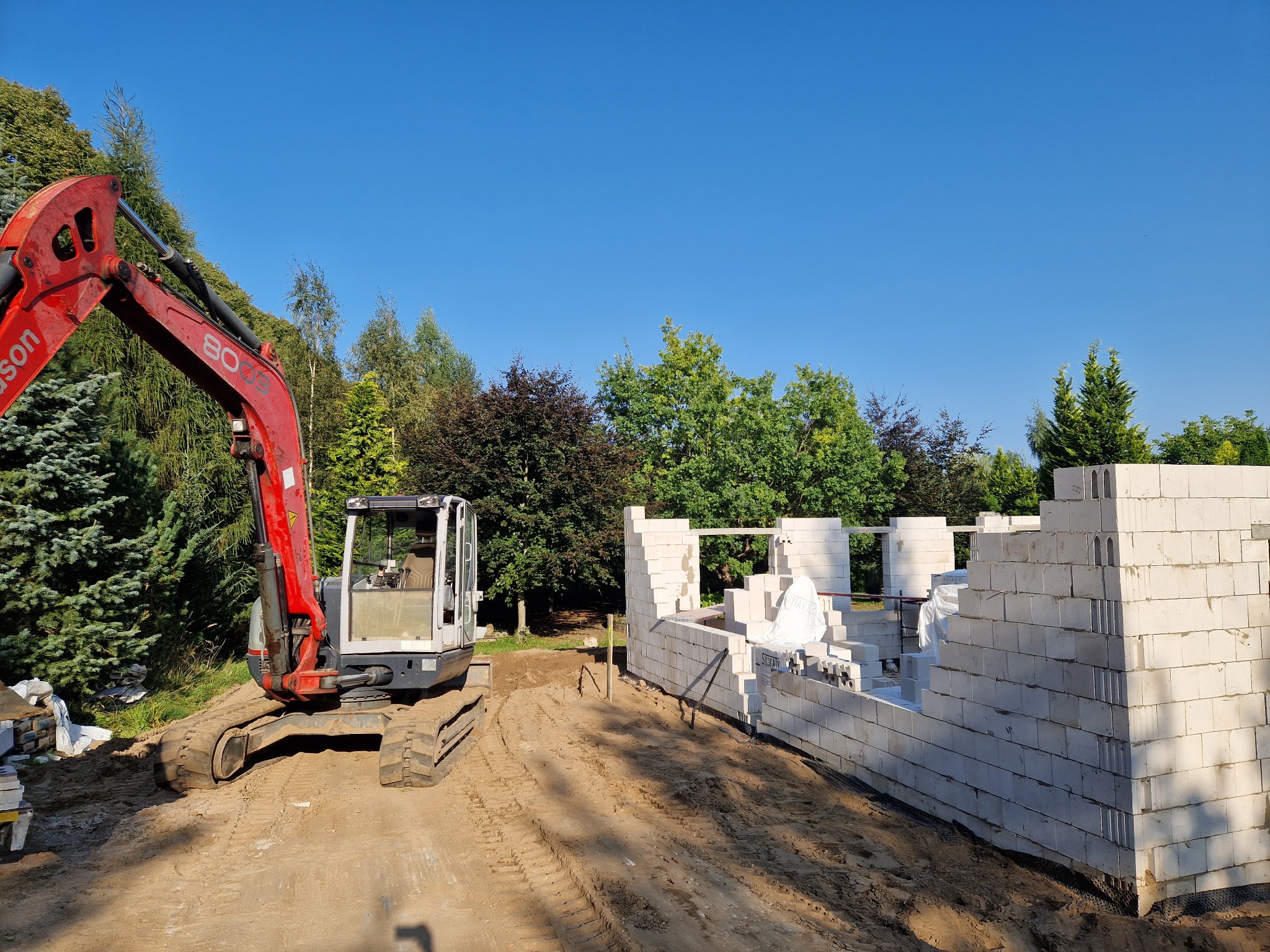 Wznoszenie ścian fundamentowych z bloczków silikatowych na fundamencie, obok stoi czerwona koparka, w tle drzewa i błękitne niebo.