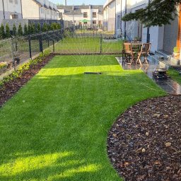 Łucki Garden Bruk - Nawadnianie trawnika z automatycznym zraszaczem na tle nowoczesnego domu jednorodzinnego z drewnianymi meblami ogrodowymi.