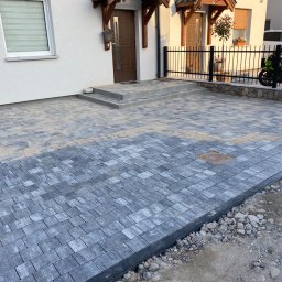 Łucki Garden Bruk - Nowa, szara kostka brukowa ułożona na podjeździe przed domem z dwoma wejściami. Widoczne schody z kostki i czarna, metalowa barierka. Częściowo podsypane piaskiem.