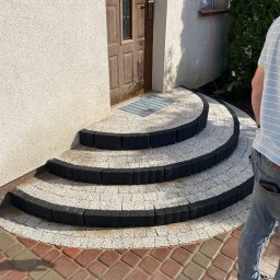 Łucki Garden Bruk - Półokrągłe, trójstopniowe schody z jasnej kostki brukowej z czarnym obrzeżem prowadzą do drzwi wejściowych. Widoczne fragmenty chodnika z czerwonej kostki i elewacji budynku.