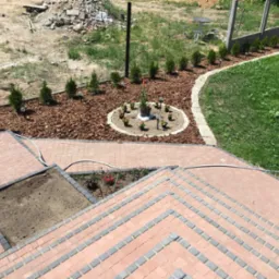 Ogród z kostką brukową, rabatami z korą i roślinami. Widoczne schody z kostki, okrągła rabata z kwiatami i lampkami, rząd małych drzewek wzdłuż ogrodzenia.