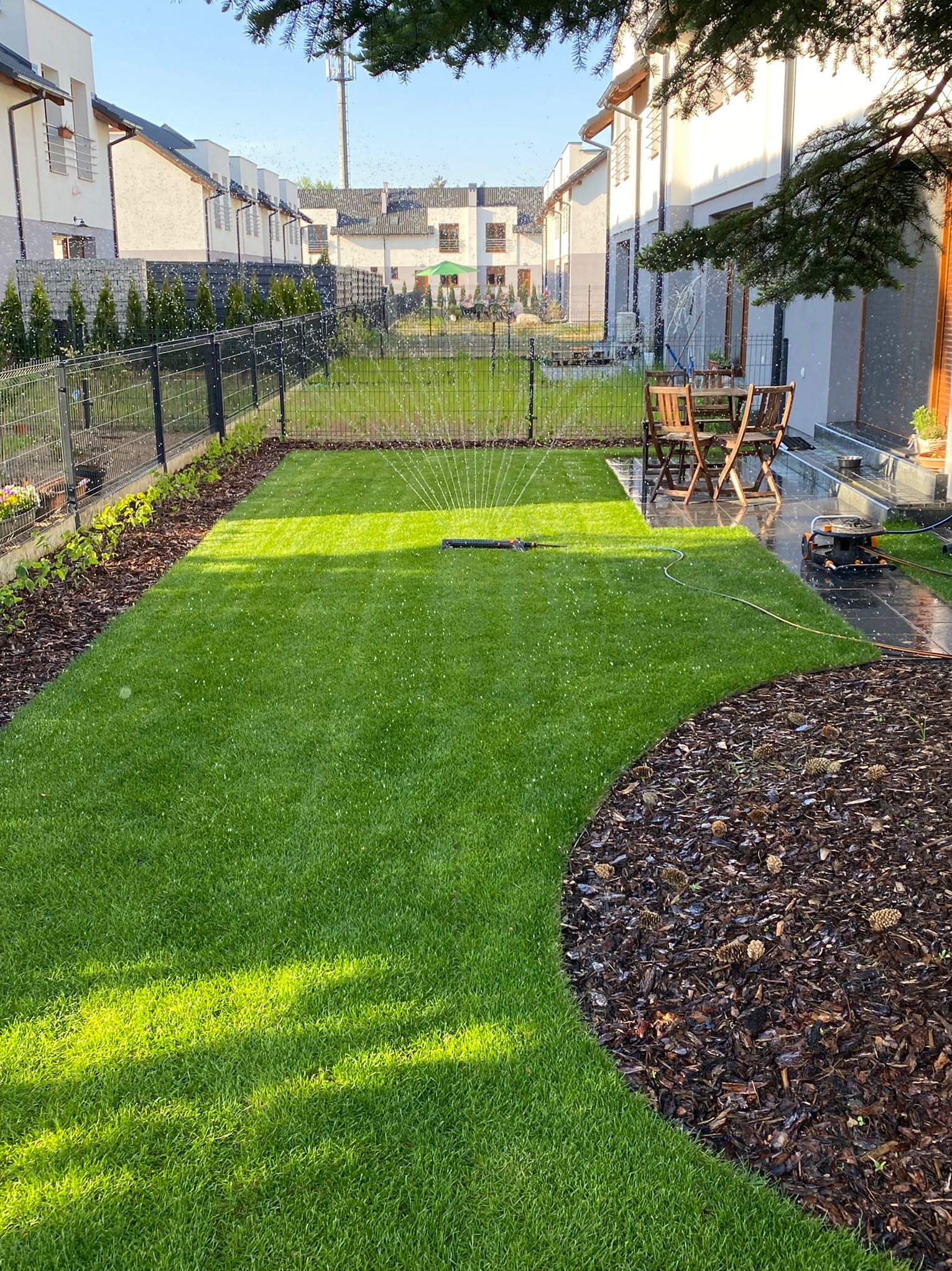 Nawadnianie trawnika z automatycznym zraszaczem na tle nowoczesnego domu jednorodzinnego z drewnianymi meblami ogrodowymi.