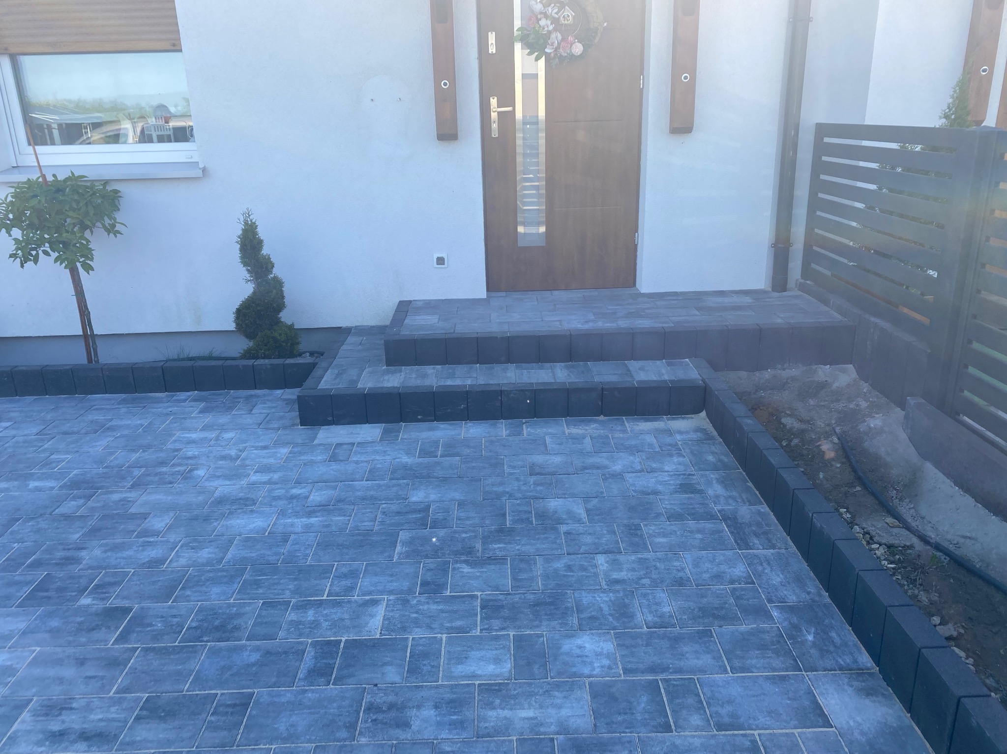 Ułożona kostka brukowa w odcieniach szarości prowadząca do wejścia domu z ozdobnym wieńcem na drzwiach, obok widoczny fragment ogrodzenia i małe drzewka ozdobne.