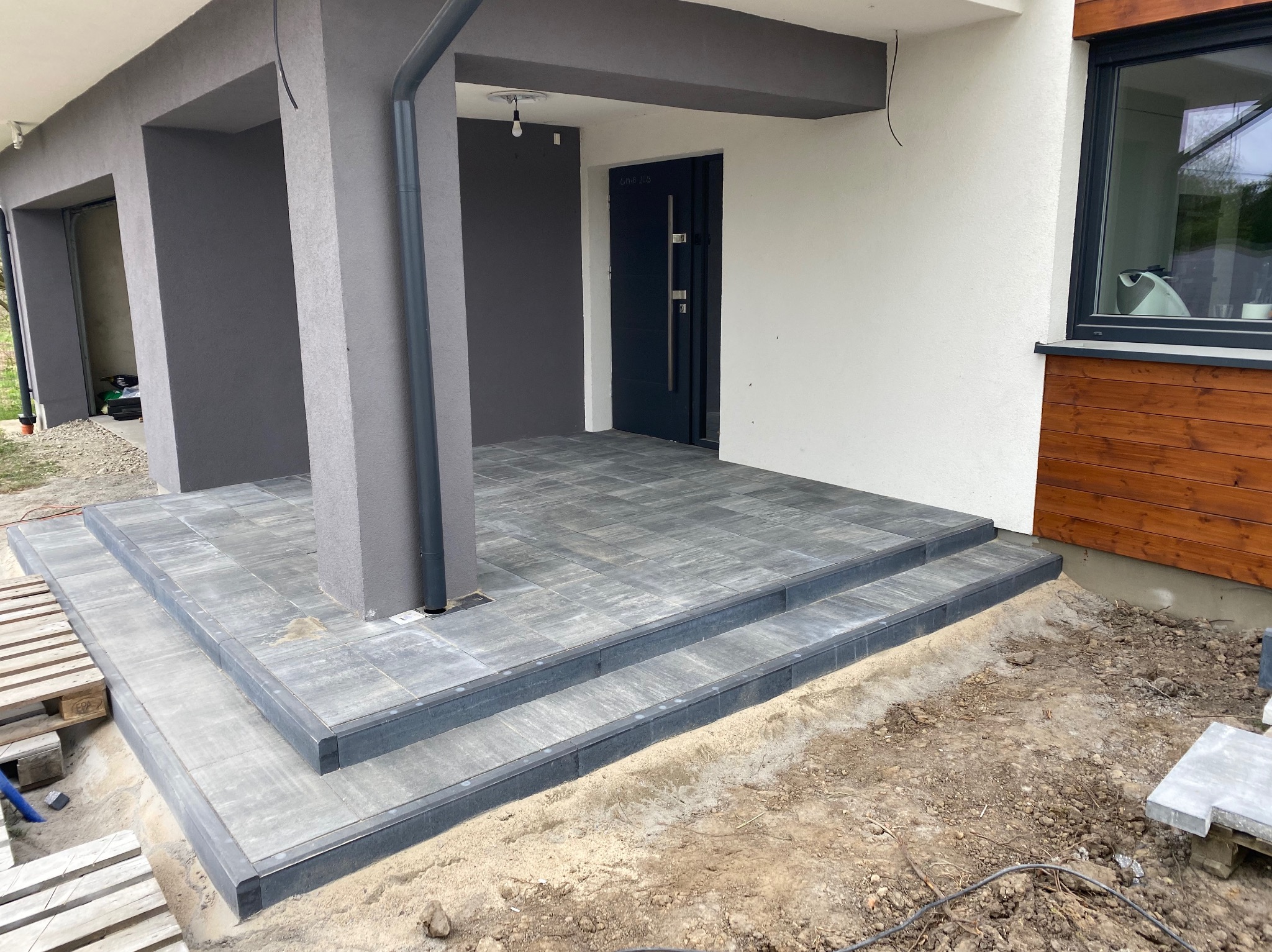 Nowoczesne wejście do domu z szarych płyt chodnikowych, dwustopniowe schody, widoczne palety i ziemia do zagospodarowania.