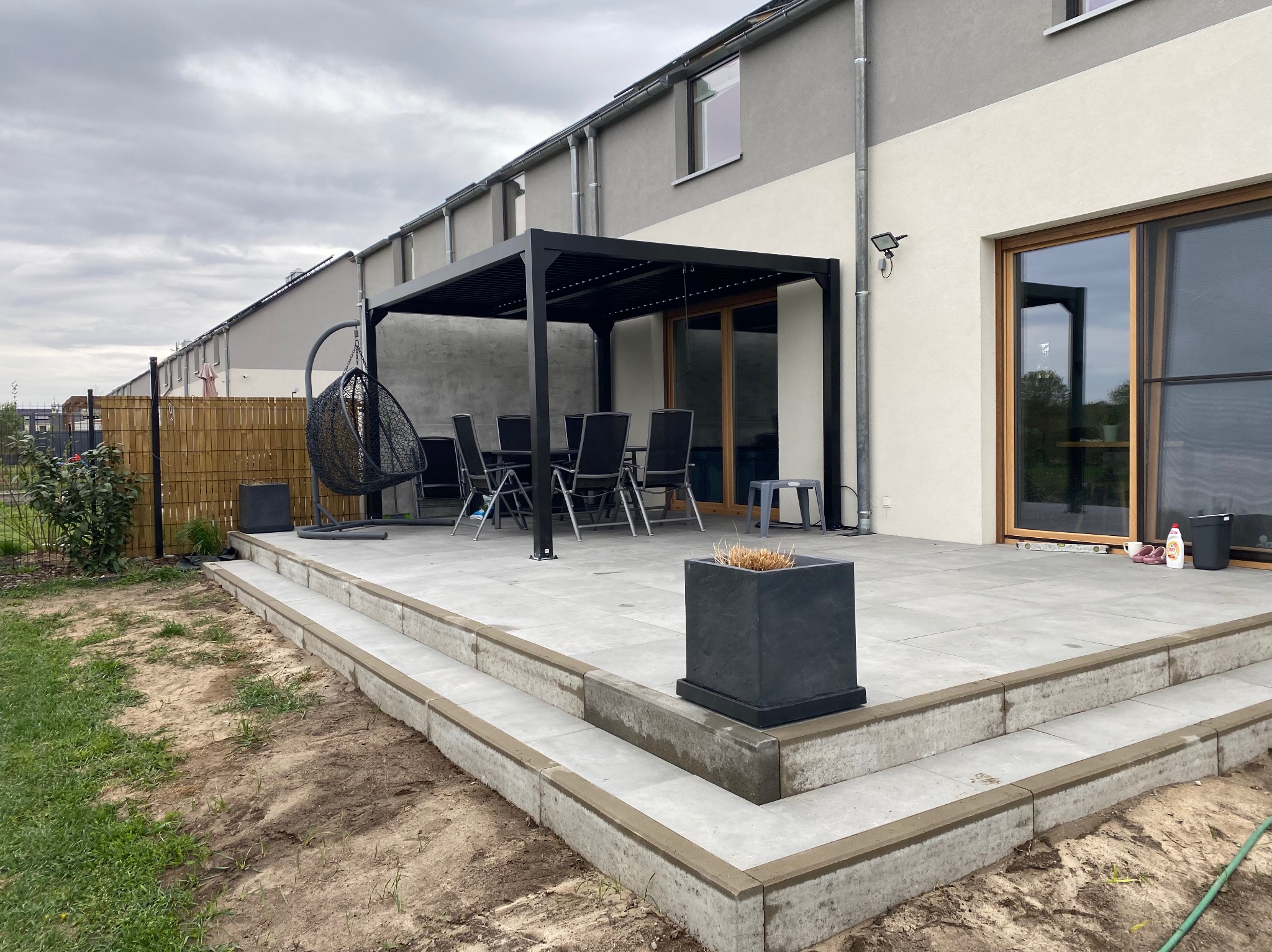 Nowoczesny taras z szarych płyt, czarna pergola z meblami ogrodowymi, wiszący fotel i donica. Schody z kostki prowadzą na taras. Tło: dom w zabudowie szeregowej.
