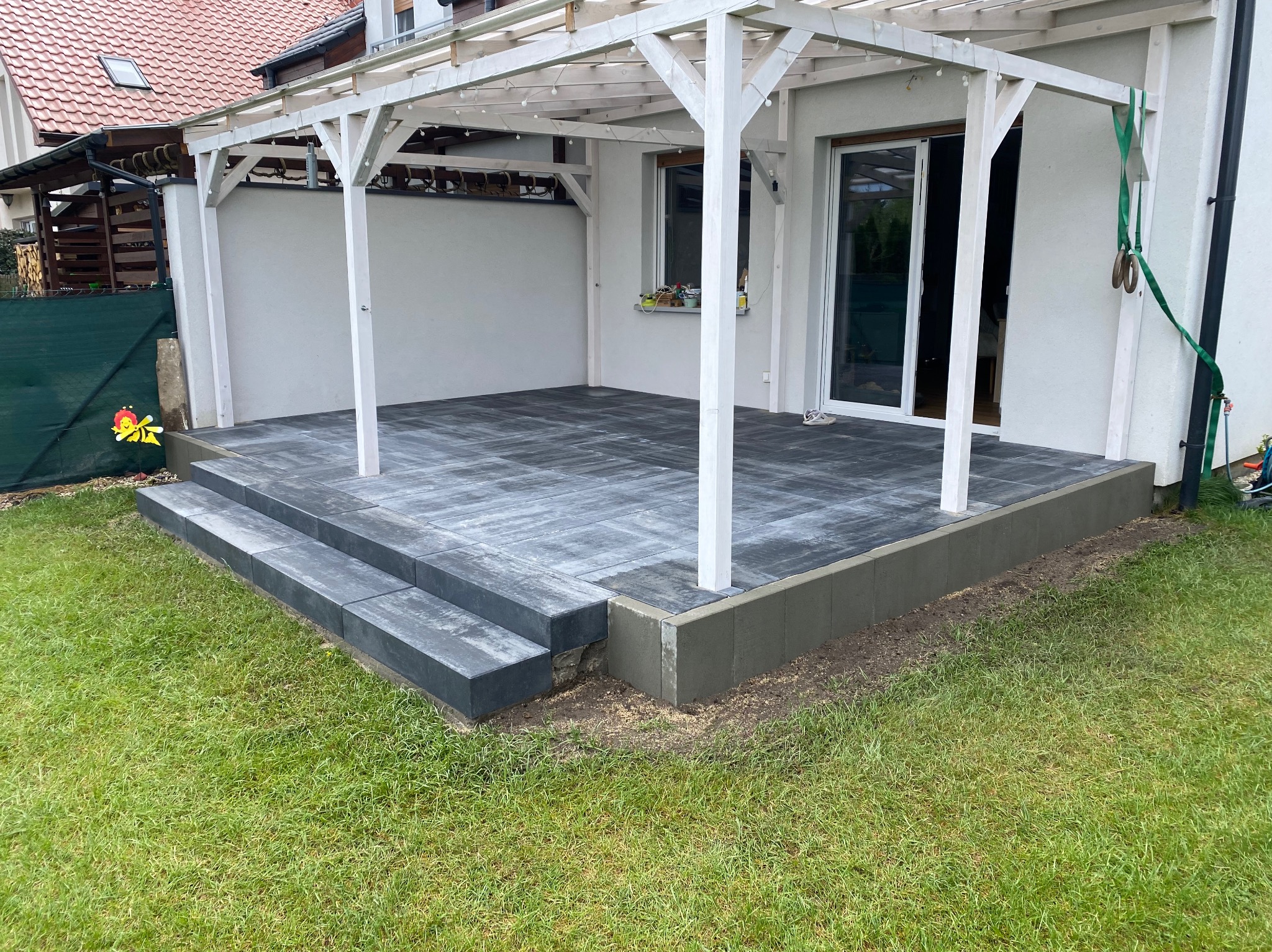 Nowoczesny taras z szarej kostki brukowej pod białą pergolą, z trzema stopniami wejściowymi. Widok na trawnik i ścianę budynku z oknem.