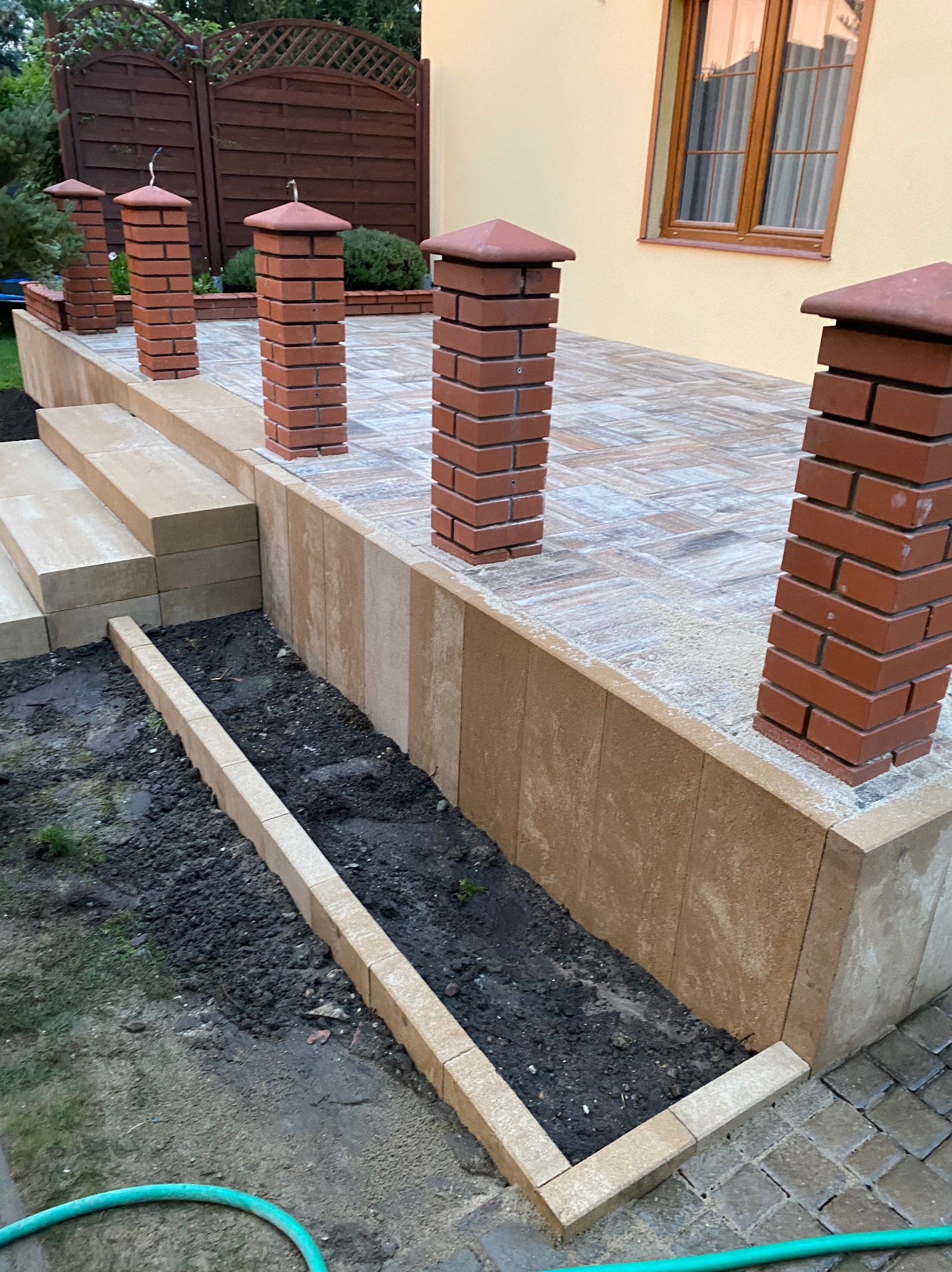 Podwyższony taras z kostki brukowej, obramowany ceglanymi słupkami i rabatą. Widoczne schody z kostki. Częściowo zagospodarowany teren.