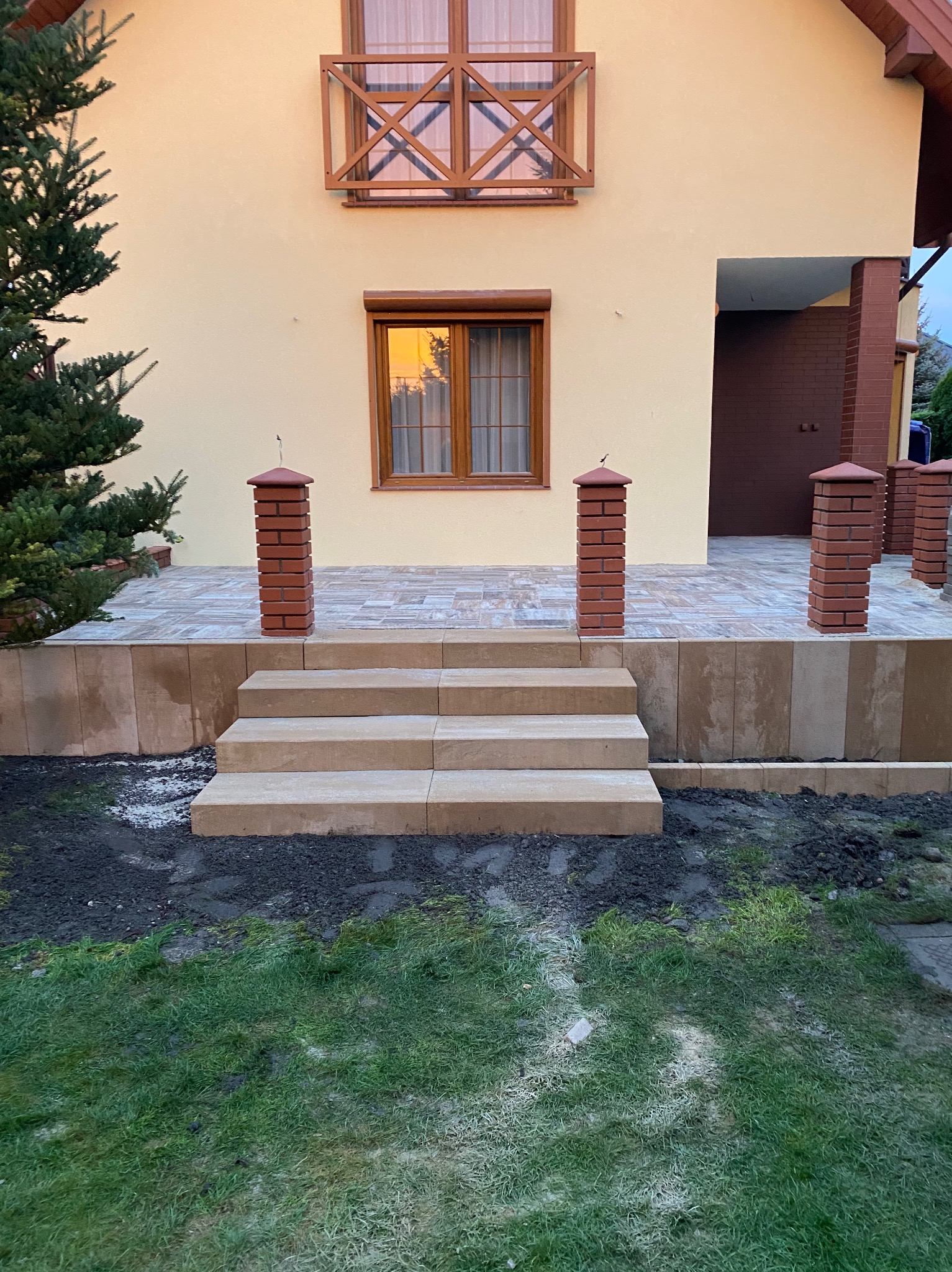 Wykończone schody wejściowe z jasnej kostki, otoczone świeżo przygotowanym gruntem pod trawnik. Czerwone, ceglane słupki dodają elegancji. Widok na elewację domu.