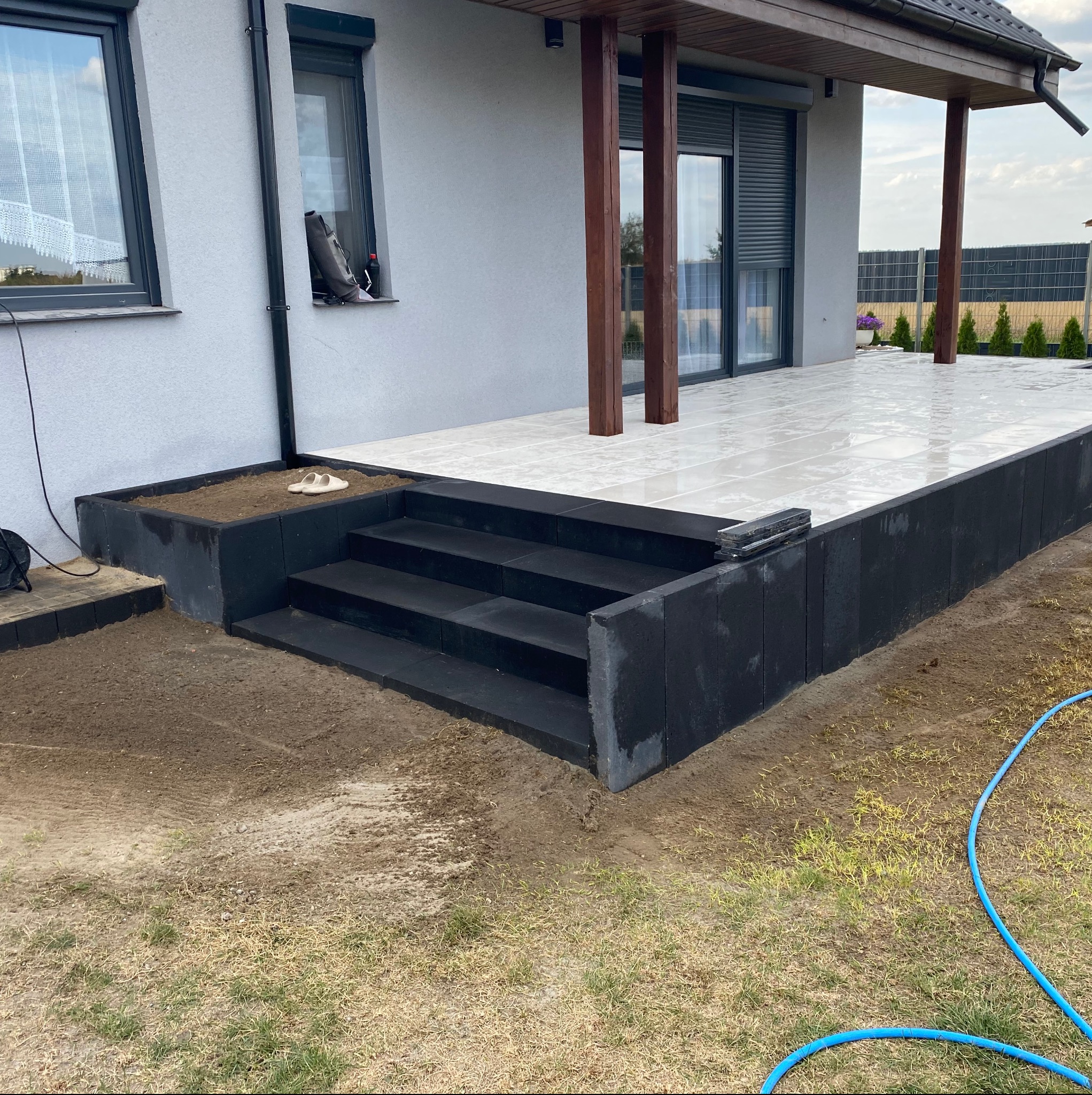 Nowoczesny taras z jasnych płyt i czarnych stopni z kostki, obok rabata z ziemią i para klapek. W tle dom z szarymi ścianami i drewnianymi elementami.