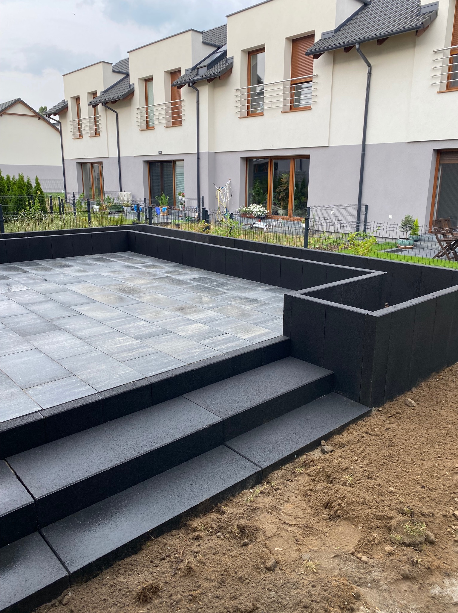 Nowoczesny taras z szarych płyt brukowych i schodami z czarnych kostek, otoczony niskim murkiem. W tle budynki mieszkalne z balkonami i zadbana zieleń.