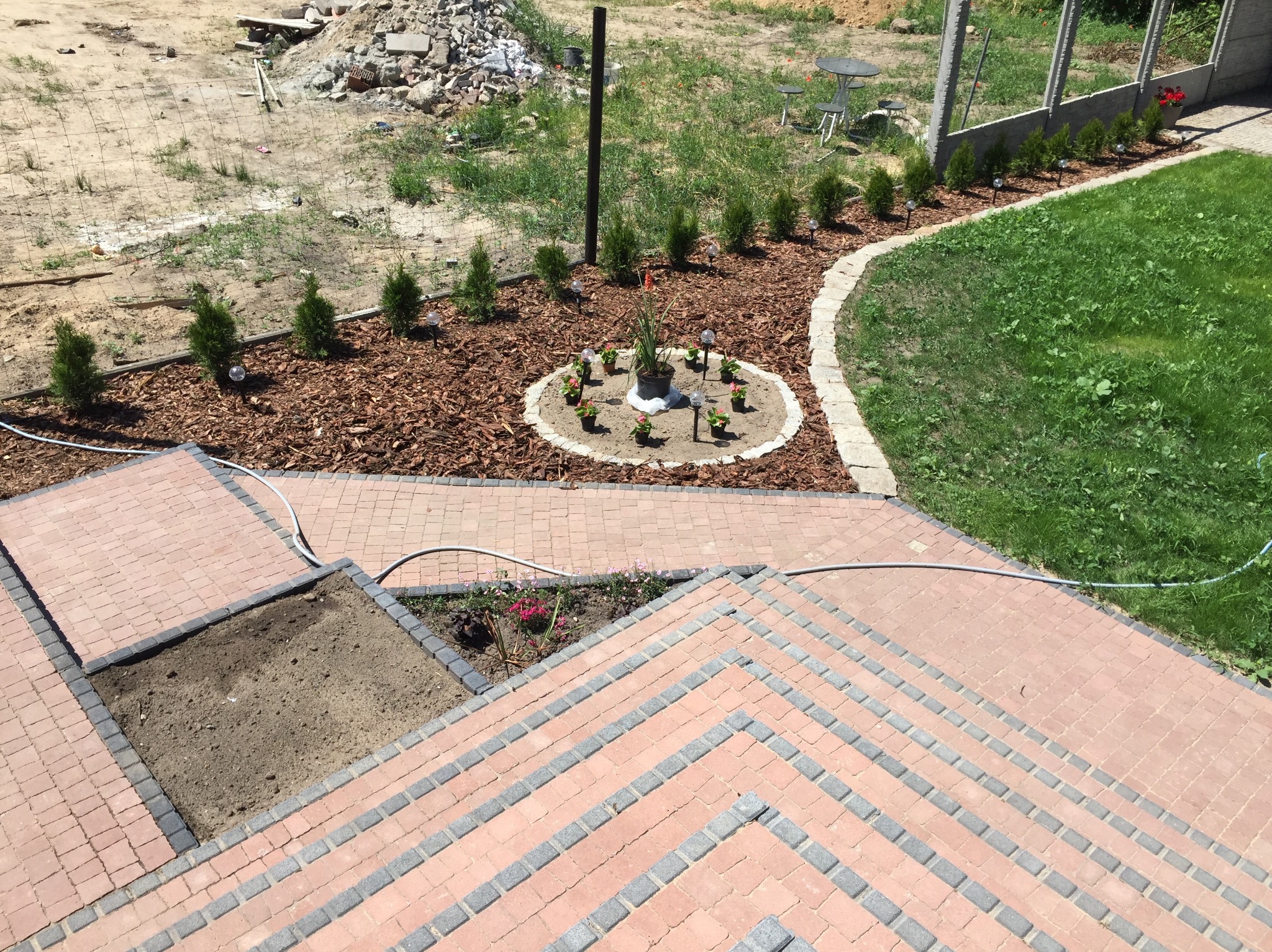 Ogród z kostką brukową, rabatami z korą i roślinami. Widoczne schody z kostki, okrągła rabata z kwiatami i lampkami, rząd małych drzewek wzdłuż ogrodzenia.