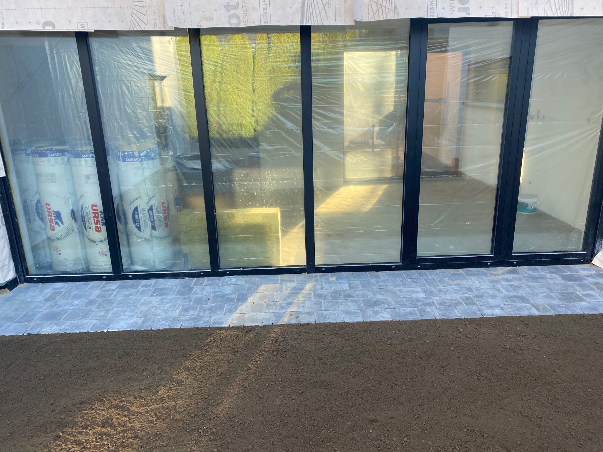 Częściowo ułożona kostka brukowa przed oknami z folią ochronną i materiałami izolacyjnymi w rolkach. Przygotowany teren pod dalsze prace.