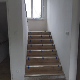 Montaż drewnopodobnych paneli na schodach z niebieską taśmą zabezpieczającą, widok z dołu, okno w tle, surowy stan ścian.