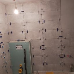 KrisBud - Ściana łazienki w trakcie układania płytek, z użyciem krzyżyków dystansowych i poziomicy. Widoczne elementy instalacji hydraulicznej i podkonstrukcja pod zabudowę.