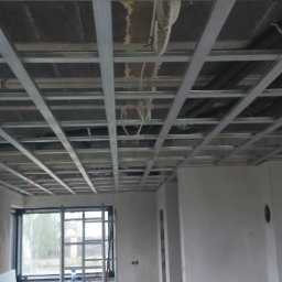 KrisBud - Sufit podwieszany z metalową konstrukcją w surowym wnętrzu. Widoczne okno z widokiem na zewnątrz. Etap prac wykończeniowych w nowym budownictwie.