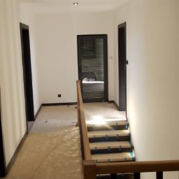 KrisBud - Korytarz w trakcie malowania, zabezpieczone schody, świeżo pomalowane ściany na biało, czarne obramowania drzwi, widoczne kable i zabezpieczenia podłogi.