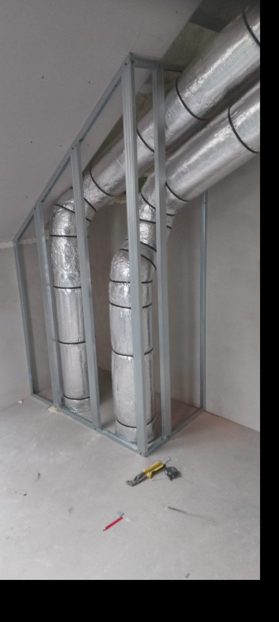 Metalowa konstrukcja z izolowanymi rurami wentylacyjnymi w surowym wnętrzu. Na podłodze leżą narzędzia: kombinerki i miarka. Widoczne niedoskonałości ścian i podłogi.