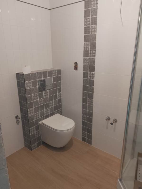 Wykończona łazienka z podwieszaną toaletą, obudową ze wzorzystych płytek i drewnopodobną podłogą. Dekoracyjny pas płytek na ścianie. Widoczny fragment kabiny prysznicowej.