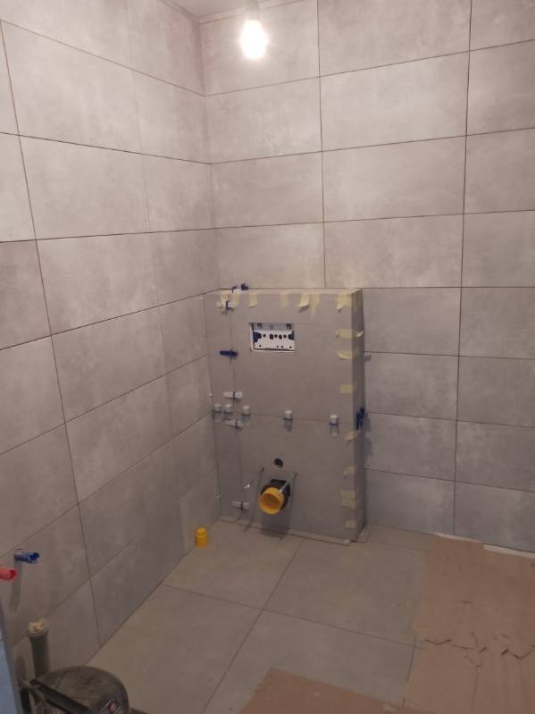 Szare, matowe płytki w łazience, zabudowa stelaża WC z widocznymi przyłączami wodno-kanalizacyjnymi. Etap prac przed ostatecznym wykończeniem.