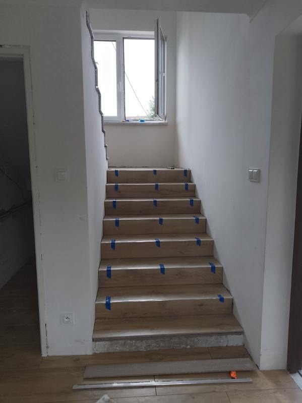 Montaż drewnopodobnych paneli na schodach z niebieską taśmą zabezpieczającą, widok z dołu, okno w tle, surowy stan ścian.