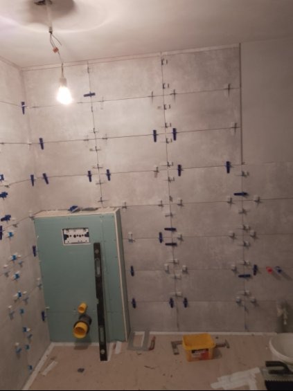 Ściana łazienki w trakcie układania płytek, z użyciem krzyżyków dystansowych i poziomicy. Widoczne elementy instalacji hydraulicznej i podkonstrukcja pod zabudowę.