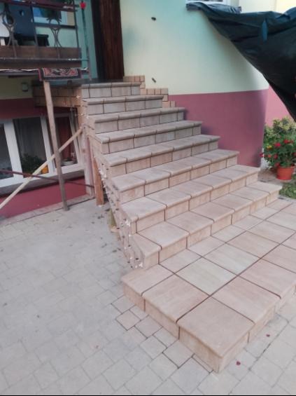 Wykończone schody zewnętrzne z beżowych płytek, prowadzące do budynku. Widoczne detale wykończenia i fuga. Częściowo widoczne rusztowanie i rośliny w tle.