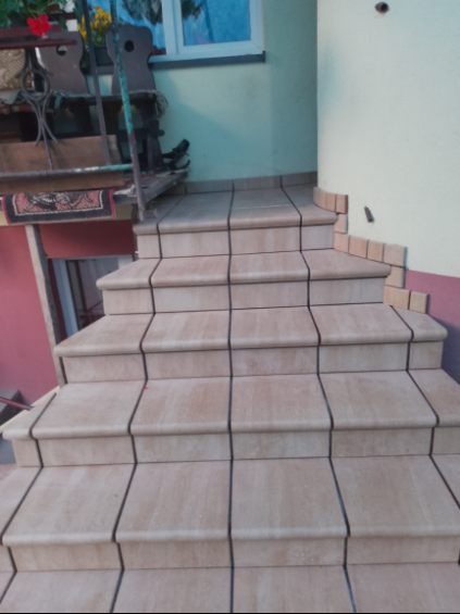 Wykończone schody zewnętrzne z beżowych płytek ceramicznych, z ciemnymi fugami, prowadzące do budynku o pastelowych ścianach. Widoczne detale wykończenia.