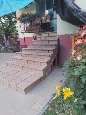 Wykończone schody zewnętrzne z beżowej kostki brukowej prowadzące do domu z balkonem, otoczone zielenią i kwiatami. Widoczne detale wykończenia stopni.