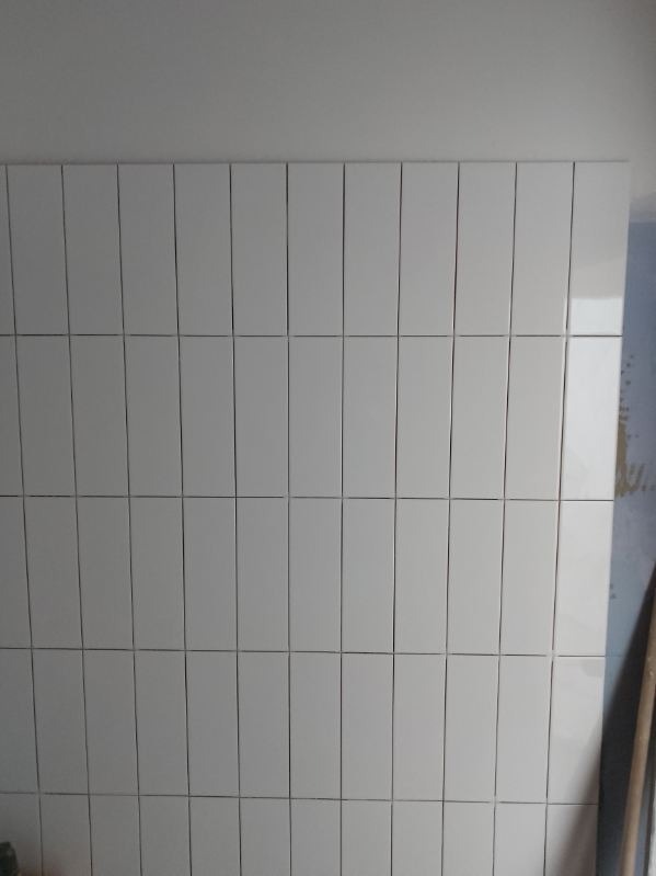 Ściana wyłożona białymi, błyszczącymi, prostokątnymi płytkami ceramicznymi ułożonymi pionowo. Widoczne fugi. Częściowo widoczny fragment ściany obok.