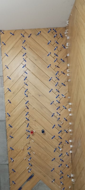 Ściana wyłożona drewnopodobnymi płytkami w jodełkę, z użyciem niebieskich i białych krzyżyków dystansowych. Widoczne otwory na instalacje. W tle szara ściana.