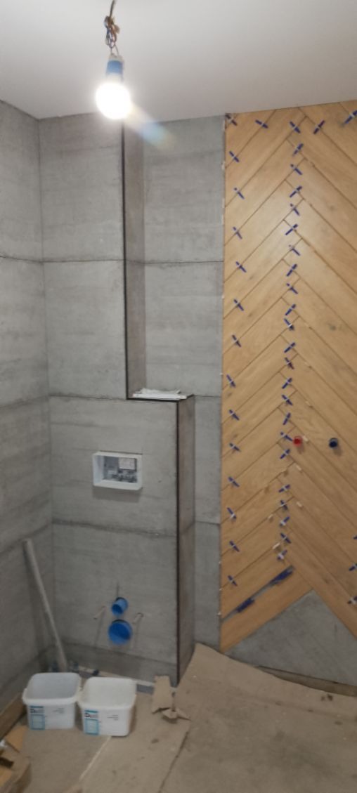 Montaż płytek drewnopodobnych w jodełkę na ścianie łazienki. Widoczne krzyżyki dystansowe i szare płytki na pozostałych ścianach. Ujęcie z dołu, sufit z żarówką.