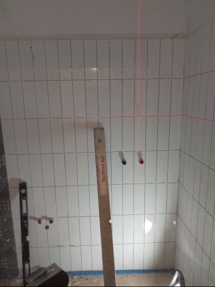 Białe, pionowo ułożone płytki w trakcie montażu w łazience. Widoczna poziomica laserowa, pionownica i fragment podłogi. Ujęcie z bliska, ukazujące etap prac glazurniczych.