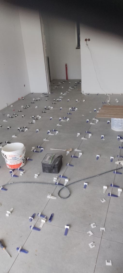 Szare płytki podłogowe w trakcie układania, z systemem poziomowania płytek. Widoczny biały pojemnik z napisem 'Gładź szpachlowa'.