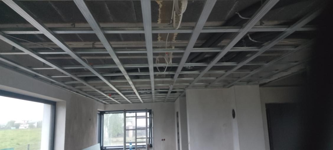 Sufit podwieszany z metalową konstrukcją w surowym wnętrzu. Widoczne okno z widokiem na zewnątrz. Etap prac wykończeniowych w nowym budownictwie.