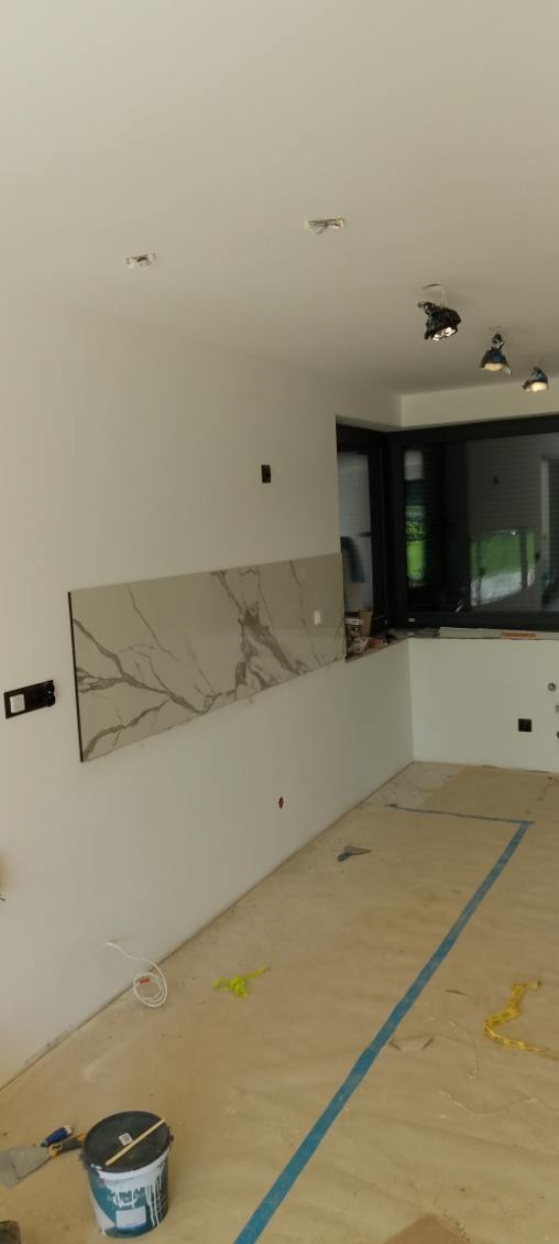 Wnętrze w trakcie wykańczania: położona długa płyta imitująca marmur na ścianie, widoczne narzędzia i wiadro z farbą na podłodze, okno w czarnej ramie.