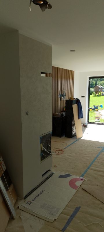 Wnętrze w trakcie wykańczania: ściana z dekoracyjną fakturą, drewniane lamele na ścianie, zabezpieczona podłoga, widok na zewnątrz. Etap prac wykończeniowych.