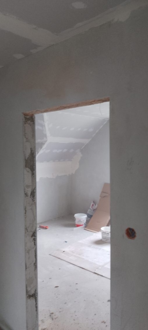 Wnętrze w trakcie prac wykończeniowych: ściany z gładzią gipsową, widoczne puszki elektryczne, fragment poddasza. Prace przygotowawcze do malowania.